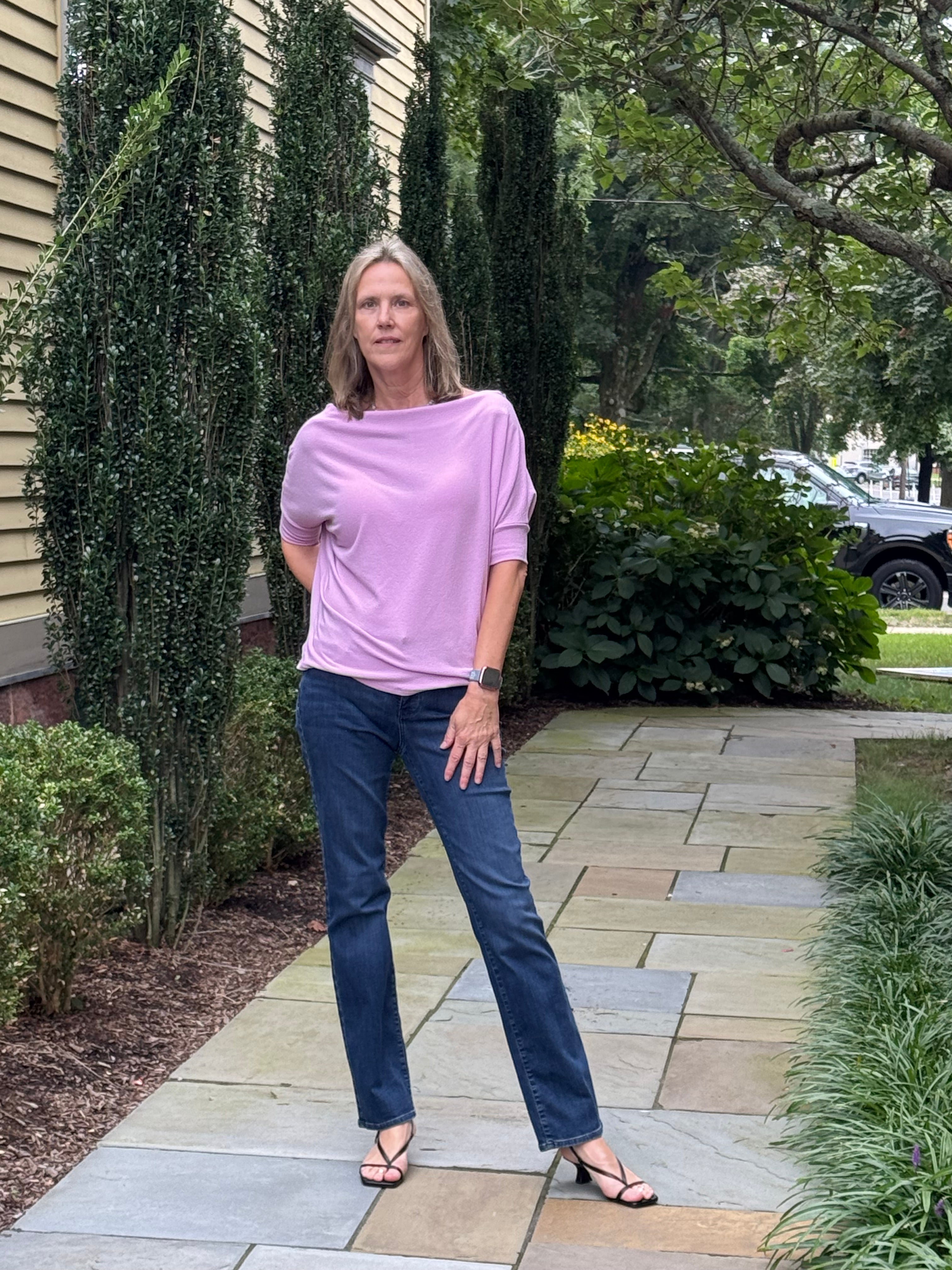 A woman stands on a stone walkway in a landscaped garden, wearing a lavender-purple boat-neck t-shirt and dark blue jeans.