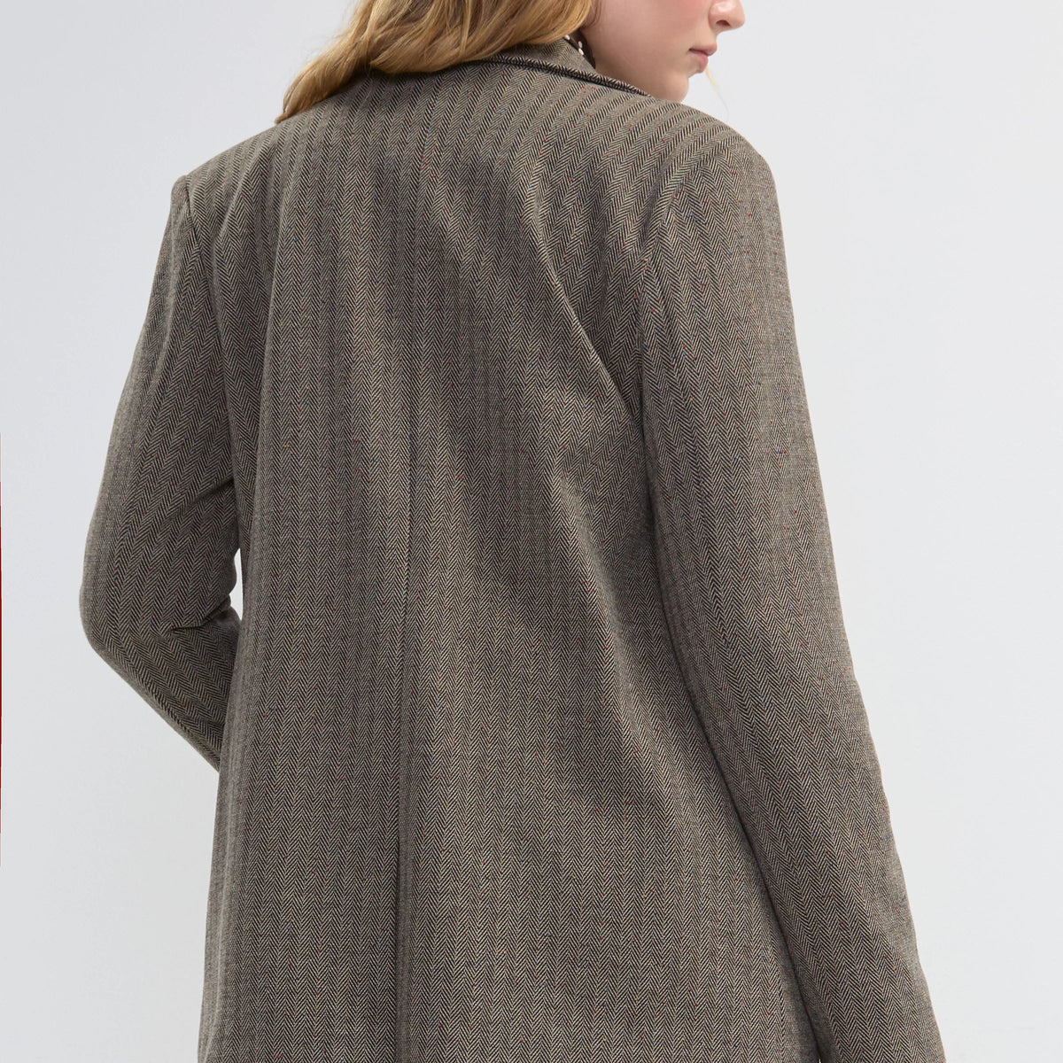 A woman shown from the back wearing a brown textured blazer with blue jeans, photographed against a light gray studio background.