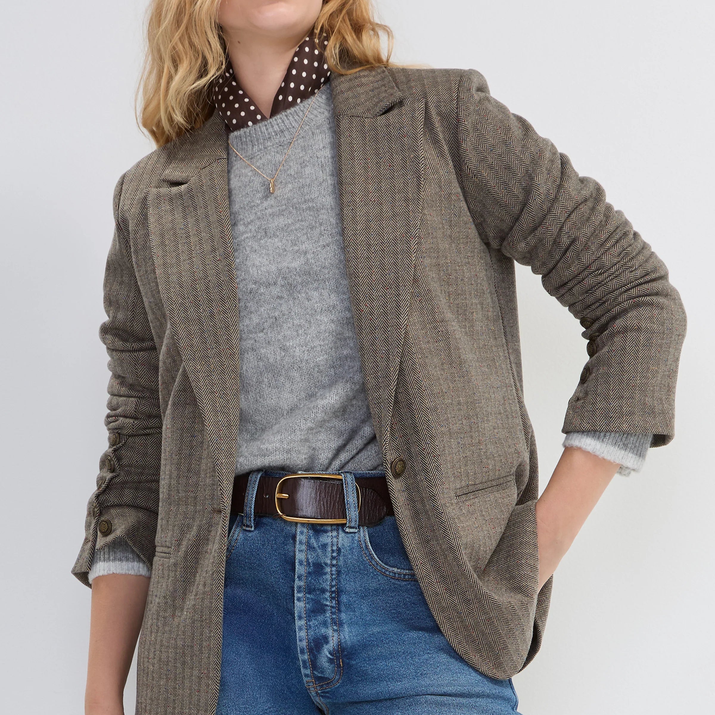 A woman modeling a brown and beige checkered blazer over a gray sweater, a polka-dot shirt collar, and blue jeans against a light gray studio background.