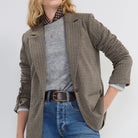 A woman modeling a brown and beige checkered blazer over a gray sweater, a polka-dot shirt collar, and blue jeans against a light gray studio background.