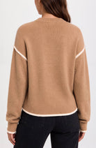 A woman viewed from the back wearing a camel-colored knit sweater with contrasting cream trim along the shoulders, hem, and cuffs, paired with dark jeans against a plain white background.