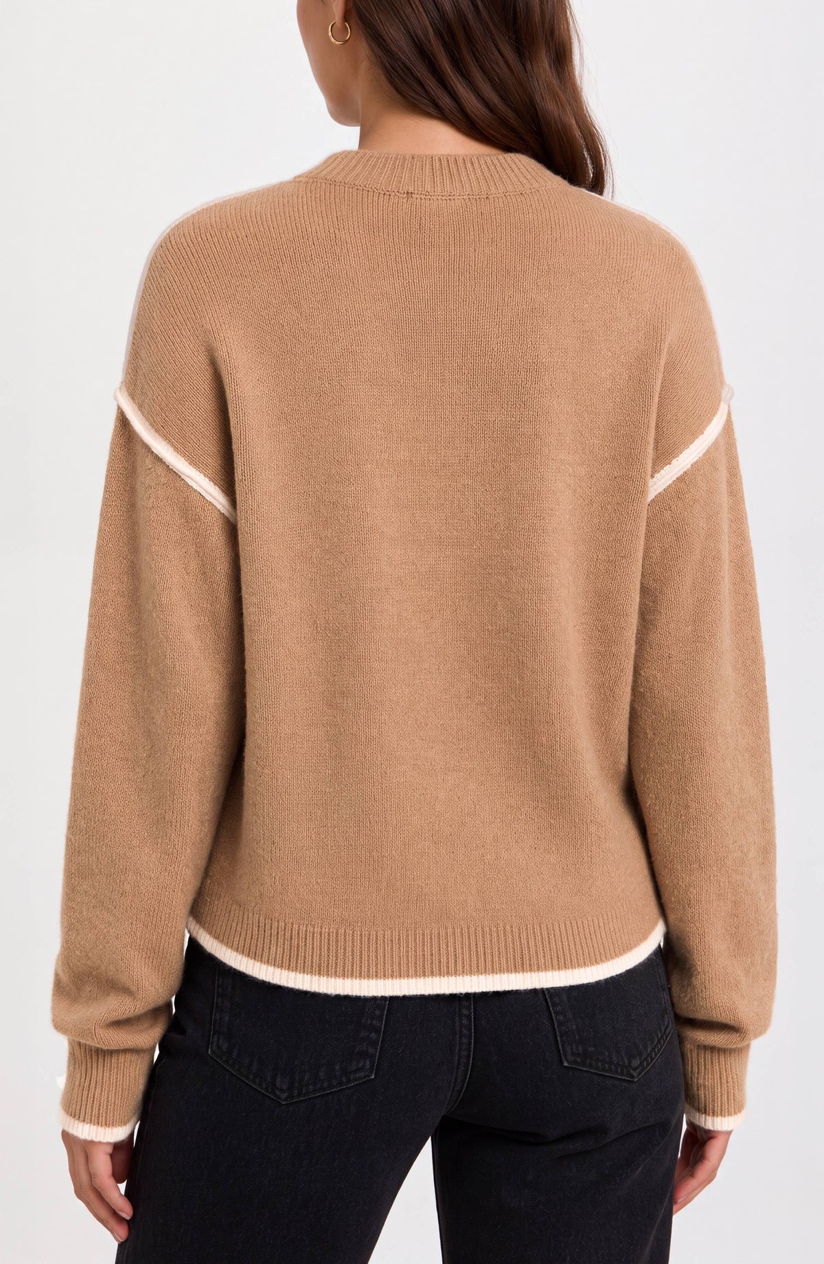 A woman viewed from the back wearing a camel-colored knit sweater with contrasting cream trim along the shoulders, hem, and cuffs, paired with dark jeans against a plain white background.