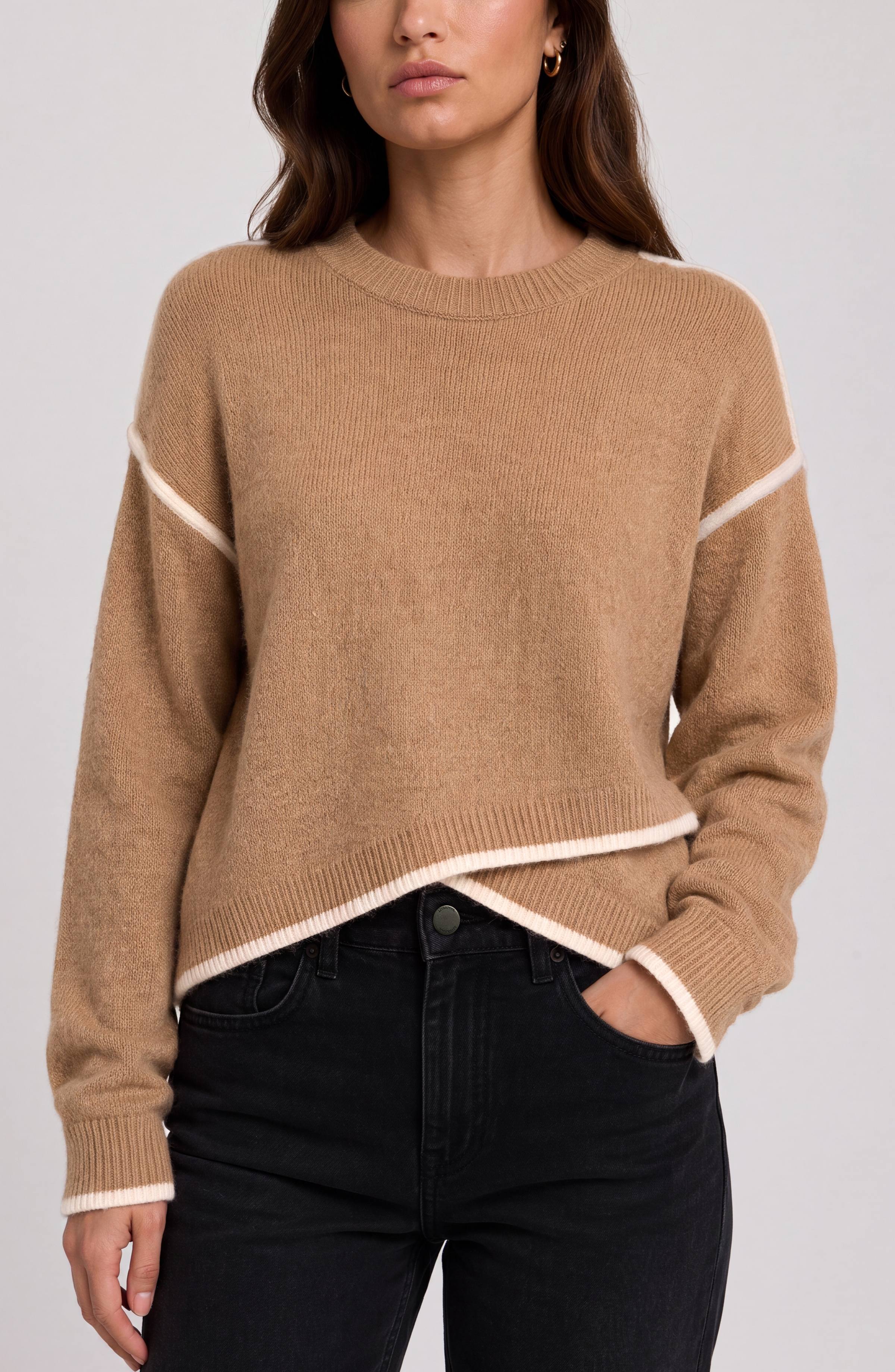 A woman wearing a cropped, long-sleeve knit sweater in camel beige with contrasting cream trim along the shoulders, hem, and cuffs, paired with black jeans against a light gray studio background.