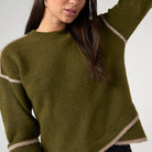 A woman wearing a fuzzy olive green knit sweater with beige contrast trim on the sleeves, collar, and hem, paired with black jeans against a light gray studio background.