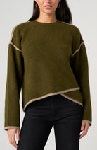A woman wearing an olive green knit sweater with beige contrast trim along the shoulders, hem, and cuffs, paired with black pants against a light gray studio background.