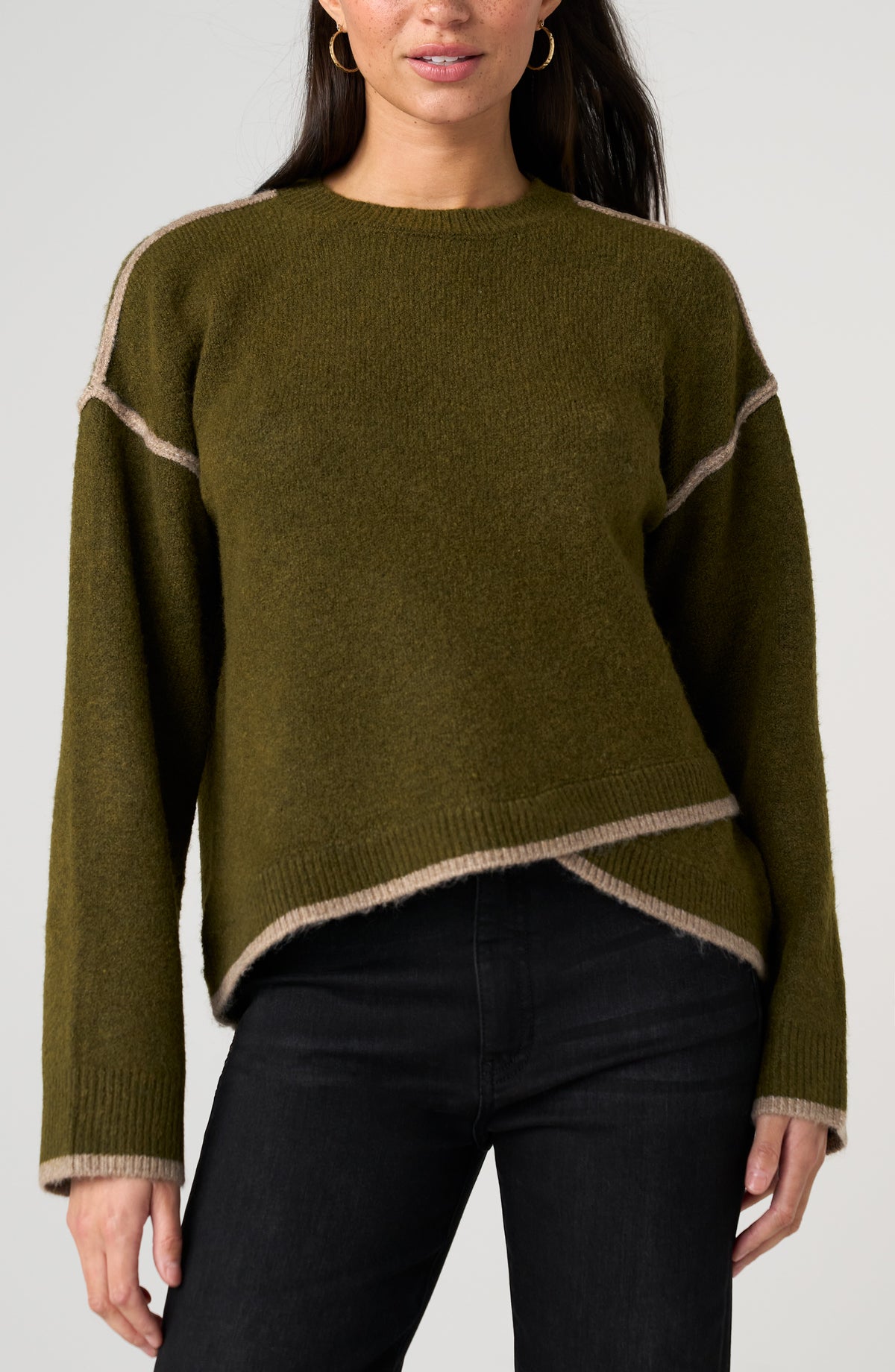 A woman wearing an olive green knit sweater with beige contrast trim along the shoulders, hem, and cuffs, paired with black pants against a light gray studio background.