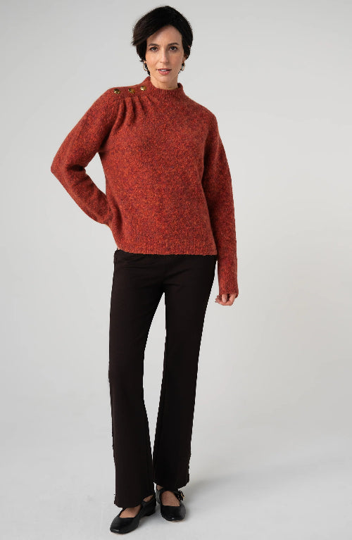 A woman modeling a textured rust-orange mock-neck knit sweater with decorative buttons on the shoulders, paired with black trousers and black flats, against a light gray studio background.