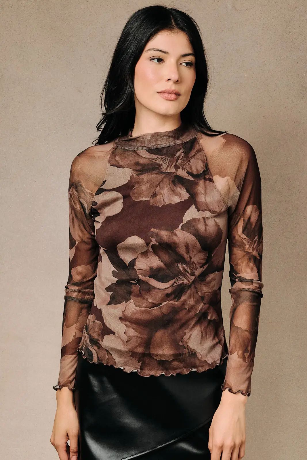 A woman wearing a sheer long-sleeve top with a brown and beige floral print, paired with a black skirt, standing against a neutral beige background.