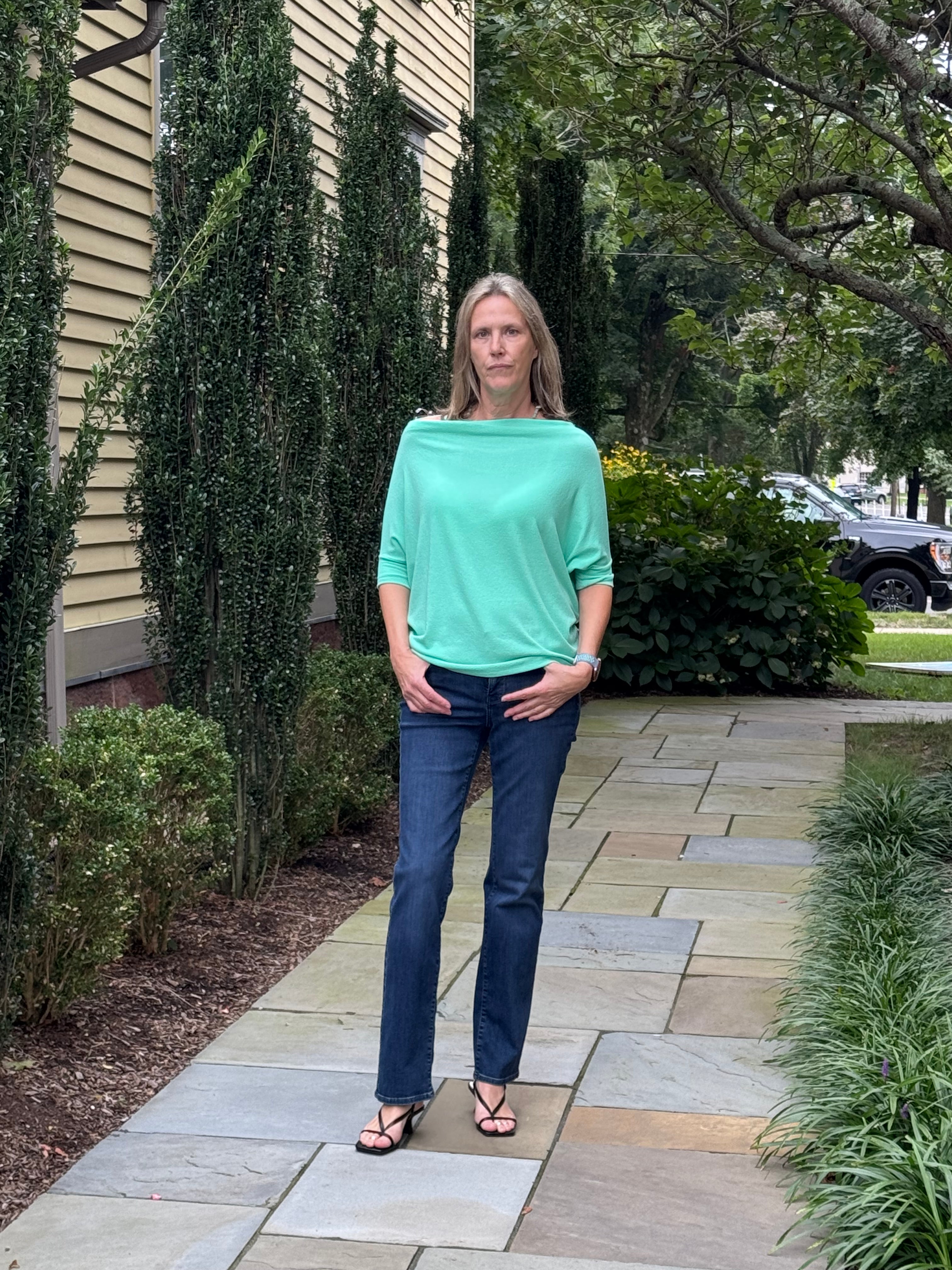 A woman stands on a stone walkway outdoors wearing a mint green off-the-shoulder top, dark blue jeans, and black sandals.