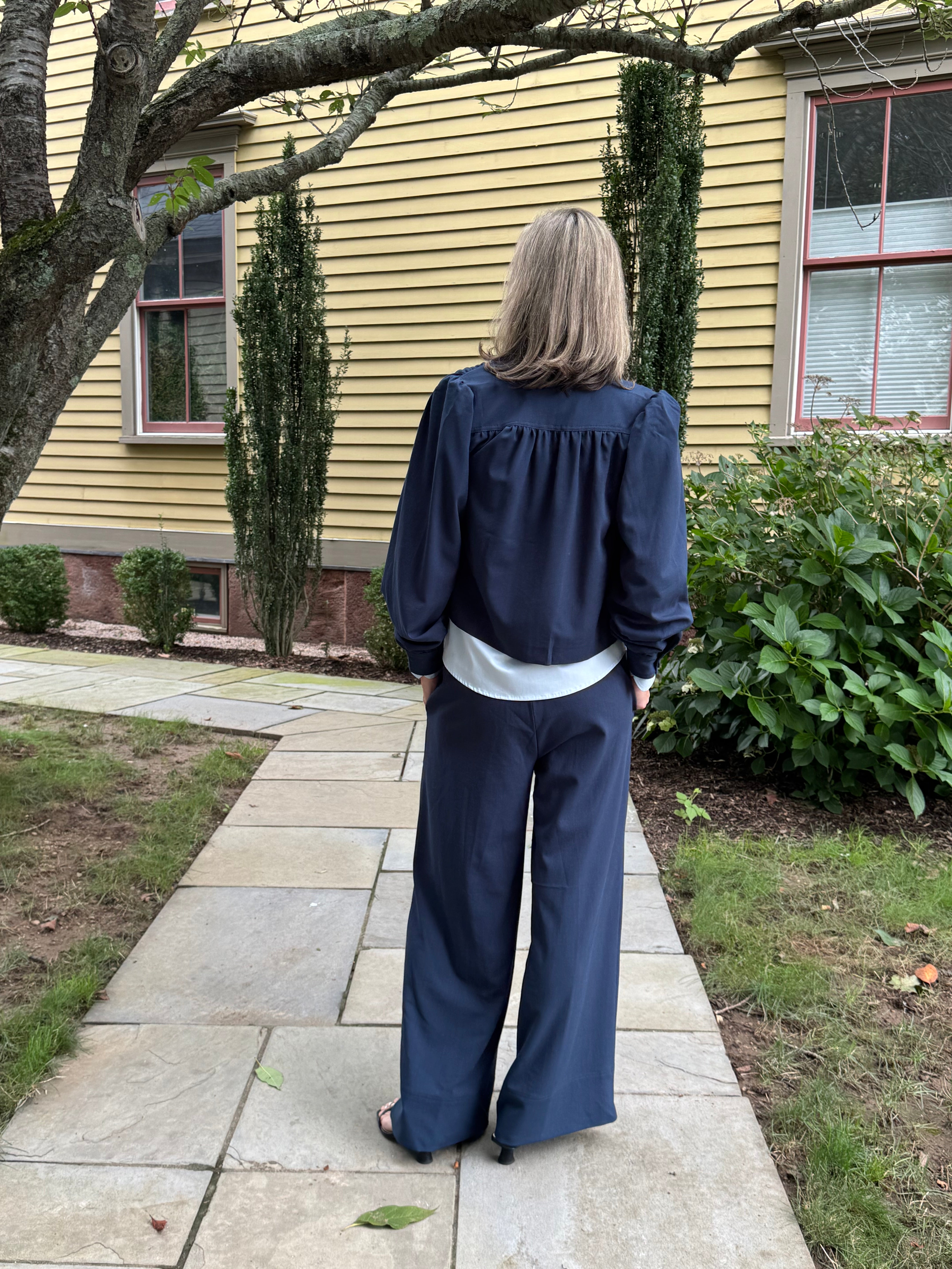 A woman stands on a stone walkway in front of a yellow house, viewed from behind. She is wearing a navy blue long-sleeve top with a gathered neckline and matching wide-leg pants.