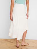 A woman wearing a flowing white midi skirt with a high-low hem, paired with a mint green top and tan flat sandals, standing in a room with light wood floors.