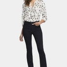A woman modeling NYDJ High Rise Billie Mini Boot Petite bootcut jeans in Huntley black wash, paired with a floral print blouse and black heels, against a plain light studio background.