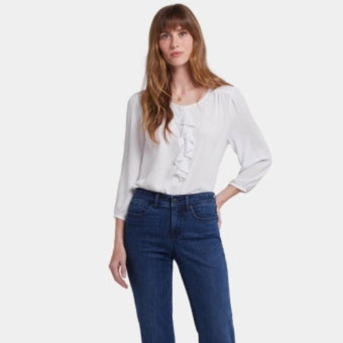 A woman modeling NYDJ Marilyn Ankle straight-leg jeans in a dark Ibiza Blues wash, paired with a white blouse and wedge sandals, against a plain light studio background.