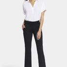 A woman modeling NYDJ Pull On Flare Ankle Trouser pants in black, paired with a white blouse, against a plain light studio background.