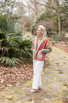A woman stands on a stone garden path wearing a colorful patchwork quilted jacket, white wide-leg pants, and sandals, with a lush garden background.