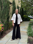A woman stands on a stone walkway outdoors in front of a house, wearing a white short-sleeve button-front jacket over a black top and black wide-leg pants.
