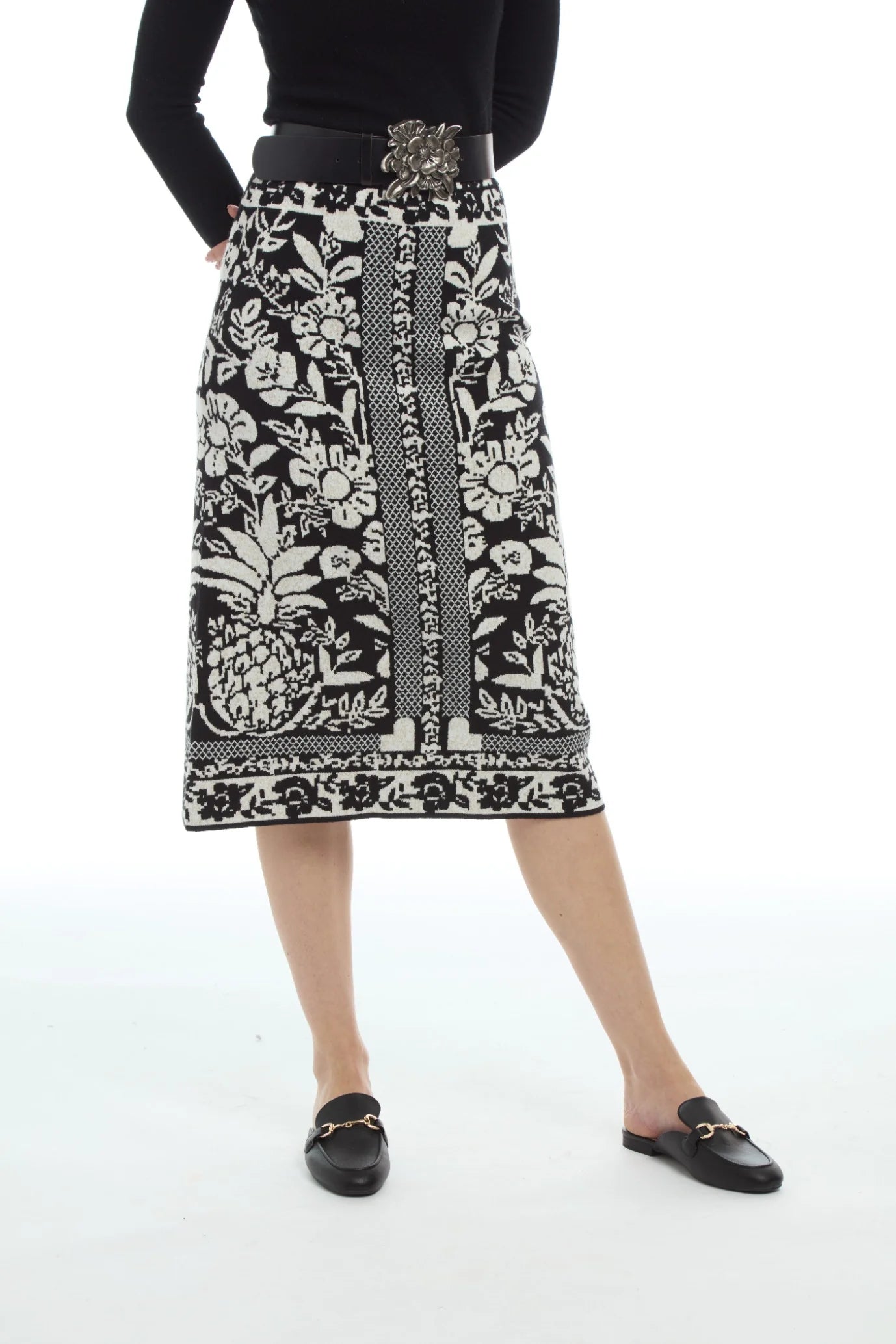 A model wearing a black and white patterned knit midi skirt with a pineapple and floral design, styled with a black top, a decorative belt, and black loafers on a white studio background.