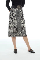 A model wearing a black and white patterned knit midi skirt with a pineapple and floral design, styled with a black top, a decorative belt, and black loafers on a white studio background.