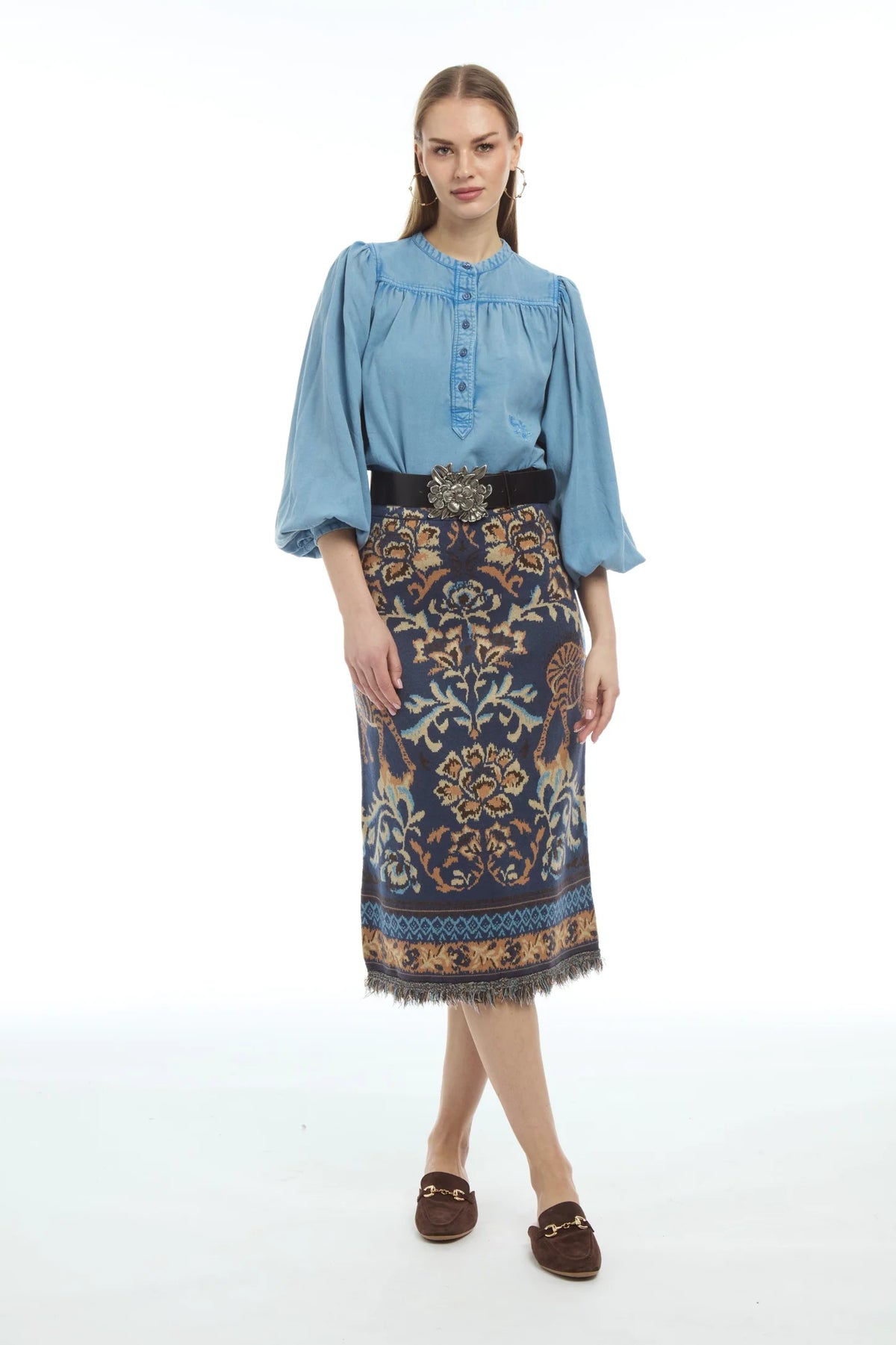 A woman modeling a light blue three-quarter sleeve blouse paired with a dark navy and gold patterned skirt and brown loafers, shot against a seamless white studio background.