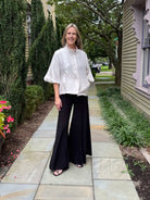 A woman stands on a stone walkway outdoors between houses, wearing a white short-sleeve button-front blouse with a high ruffled collar and black wide-leg pants.
