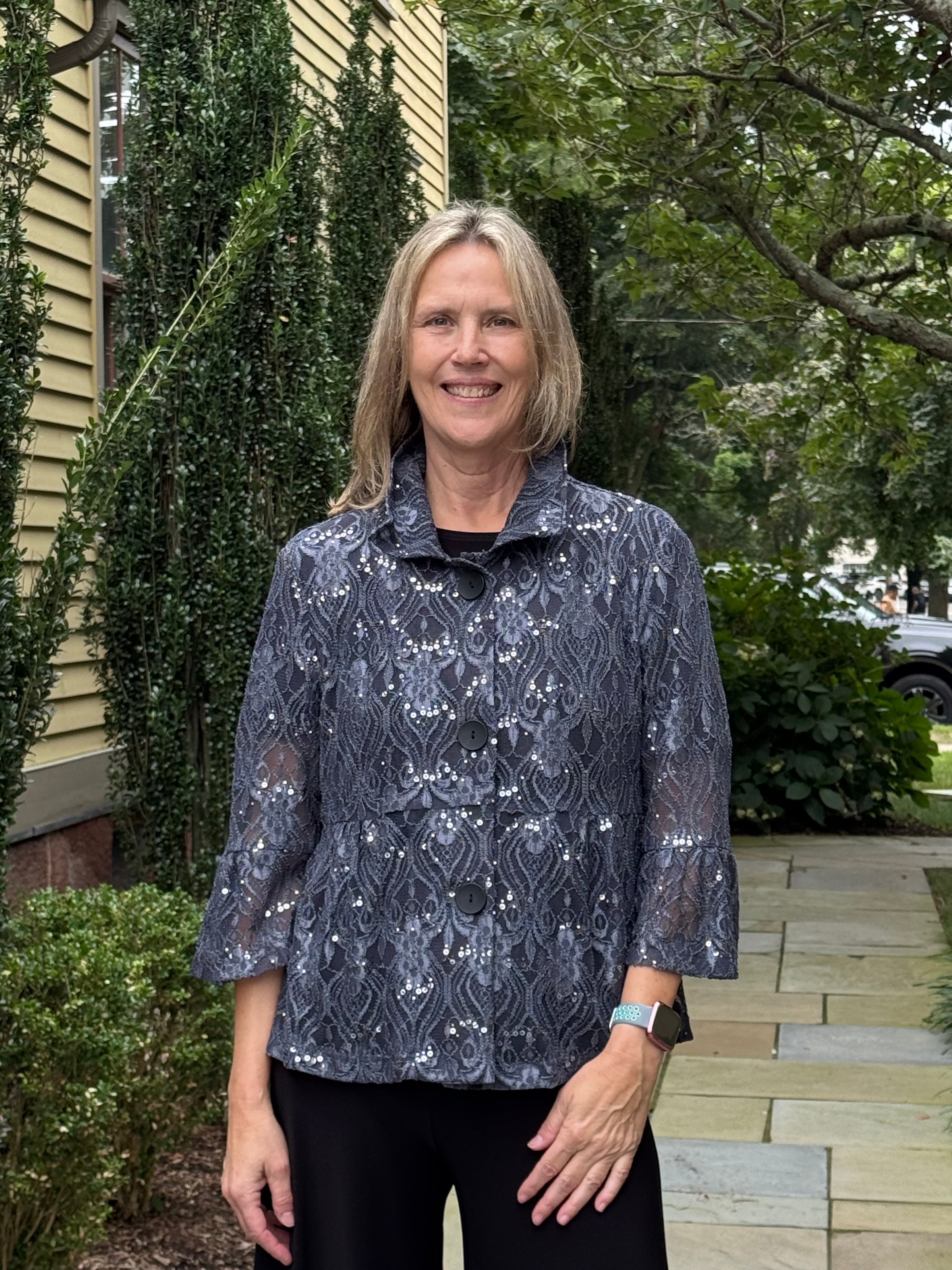 A woman with blonde hair stands outdoors on a stone walkway wearing a dark gray patterned lace top and black pants, with a house and greenery in the background.