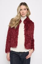 Woman wearing the Picadilly Faux Feather Short Jacket in Cabernet, a cropped plush jacket with a stand collar, over a cream mock-neck top and dark jeans