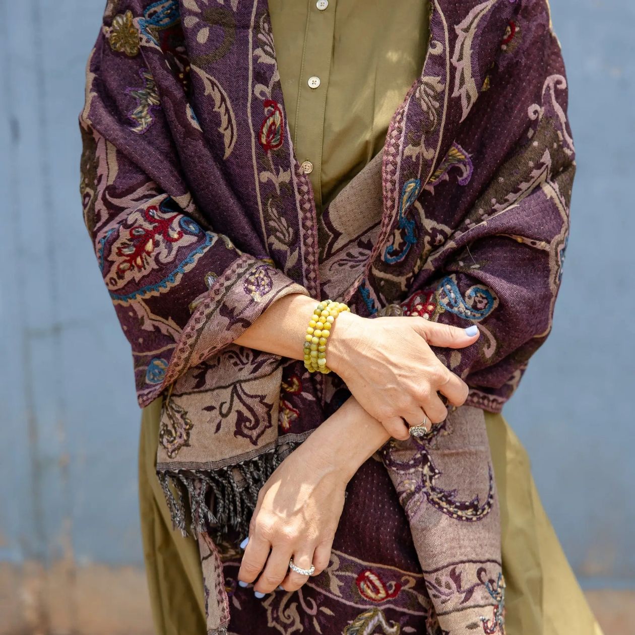 A close-up of a person wearing a patterned shawl in deep purple, beige, and brown over an olive green kurta, with yellow beaded bracelets on one wrist.