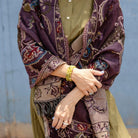 A close-up of a person wearing a patterned shawl in deep purple, beige, and brown over an olive green kurta, with yellow beaded bracelets on one wrist.