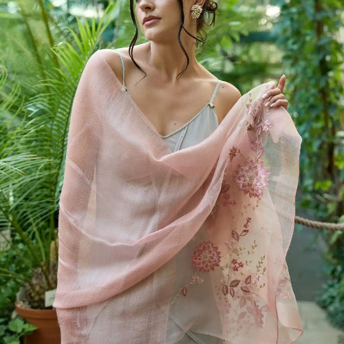 A woman wearing a SAACHI Floral Embroidered Silk Scarf, a sheer blush pink wrap with delicate floral embroidery, draped over her shoulders in a garden setting.