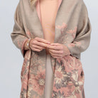 A woman wearing a reversible SAACHI wrap scarf in taupe with a pink floral pattern lining, draped open over a beige dress, against a plain light studio background.
