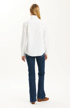 A woman viewed from the back wearing a white long-sleeve button-up shirt, dark blue flared jeans, and brown shoes against a plain white studio background.