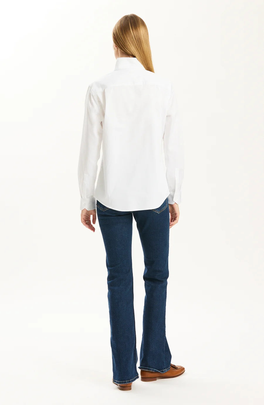 A woman viewed from the back wearing a white long-sleeve button-up shirt, dark blue flared jeans, and brown shoes against a plain white studio background.