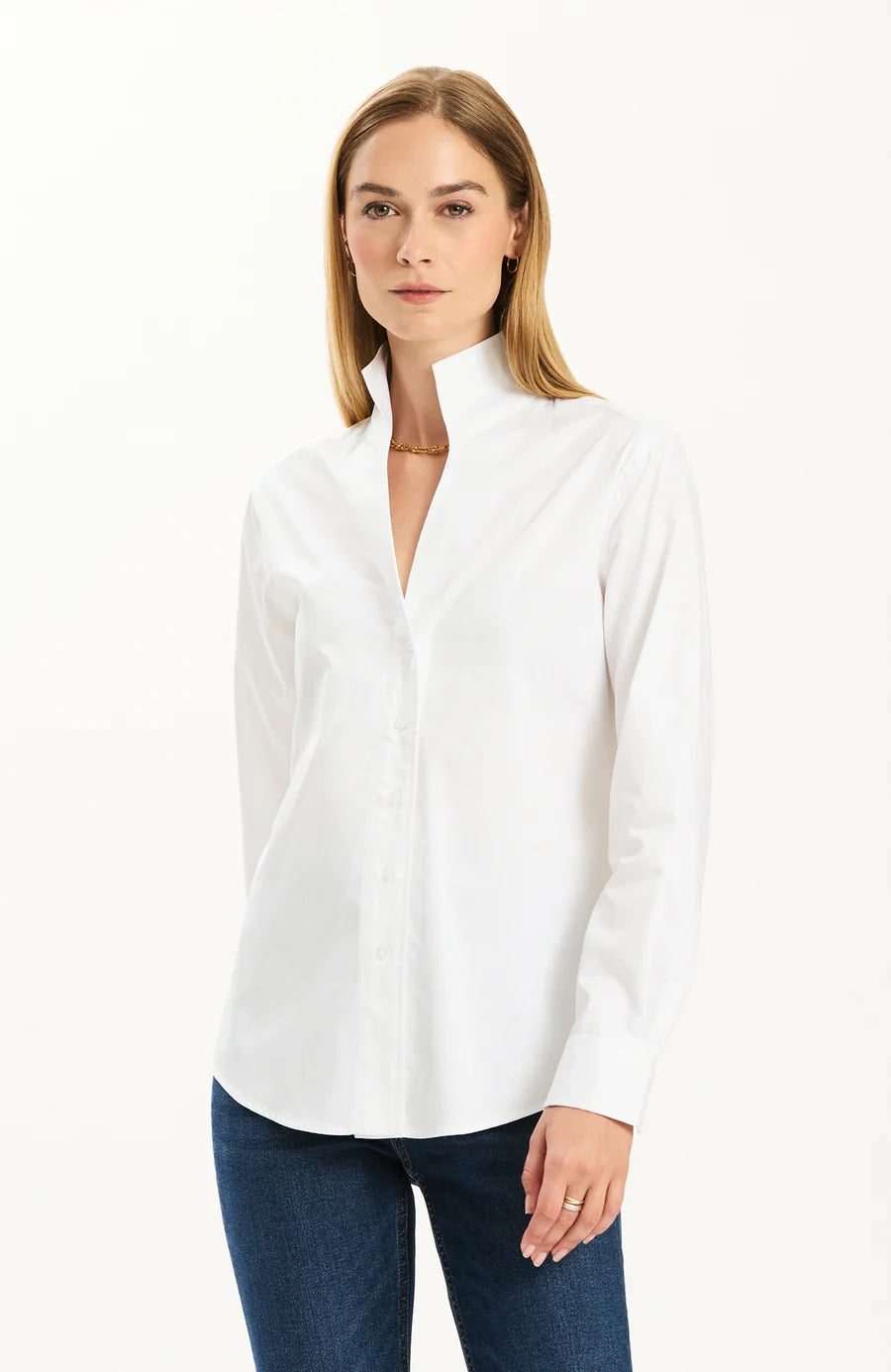 A woman wearing a white long-sleeve button-up shirt with a high stand collar and dark blue jeans, standing against a plain white studio background.