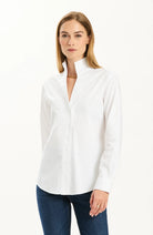 A woman wearing a white long-sleeve button-up shirt with a high stand collar and dark blue jeans, standing against a plain white studio background.