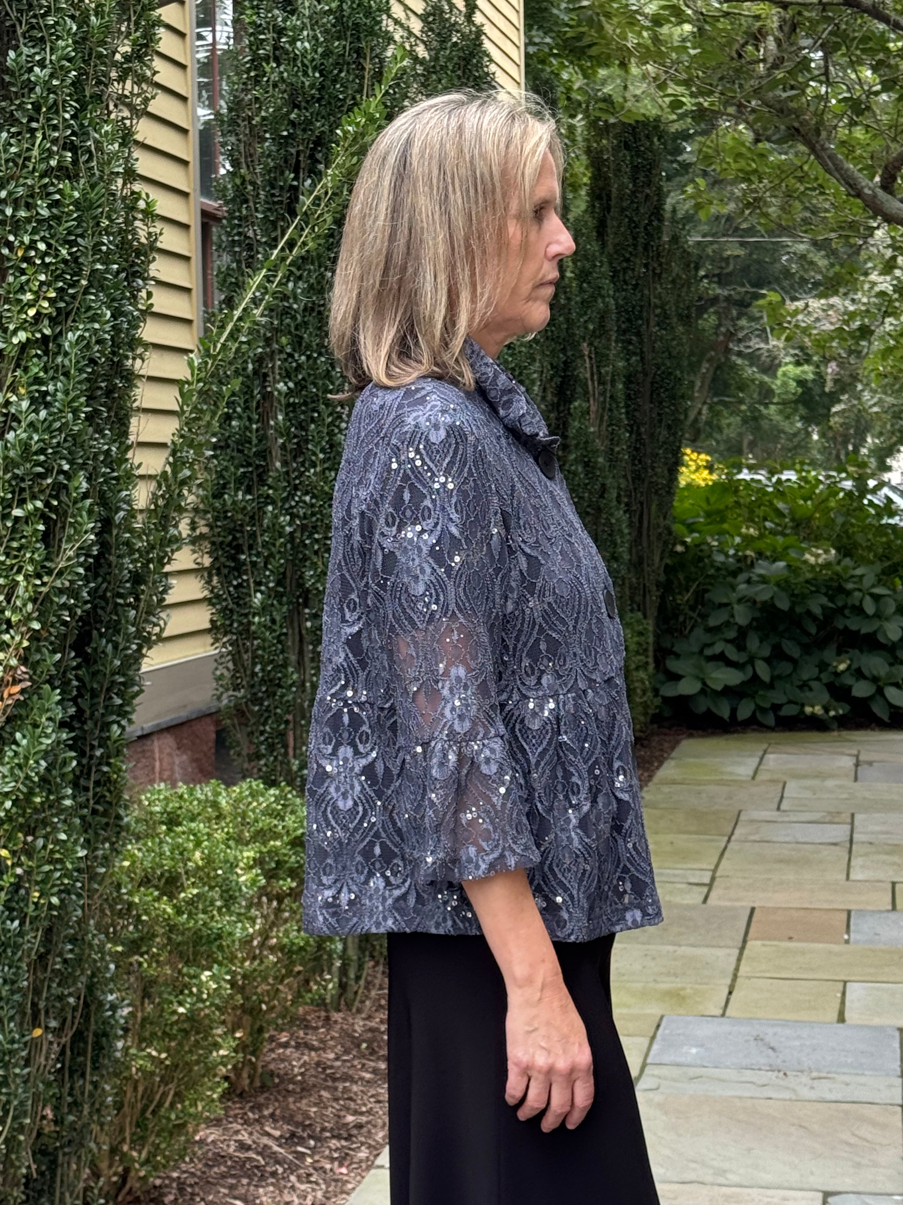 A woman standing outdoors in profile wearing a dark gray lace jacket with silver sequins over a black skirt, set against a background of green hedges and a stone walkway.