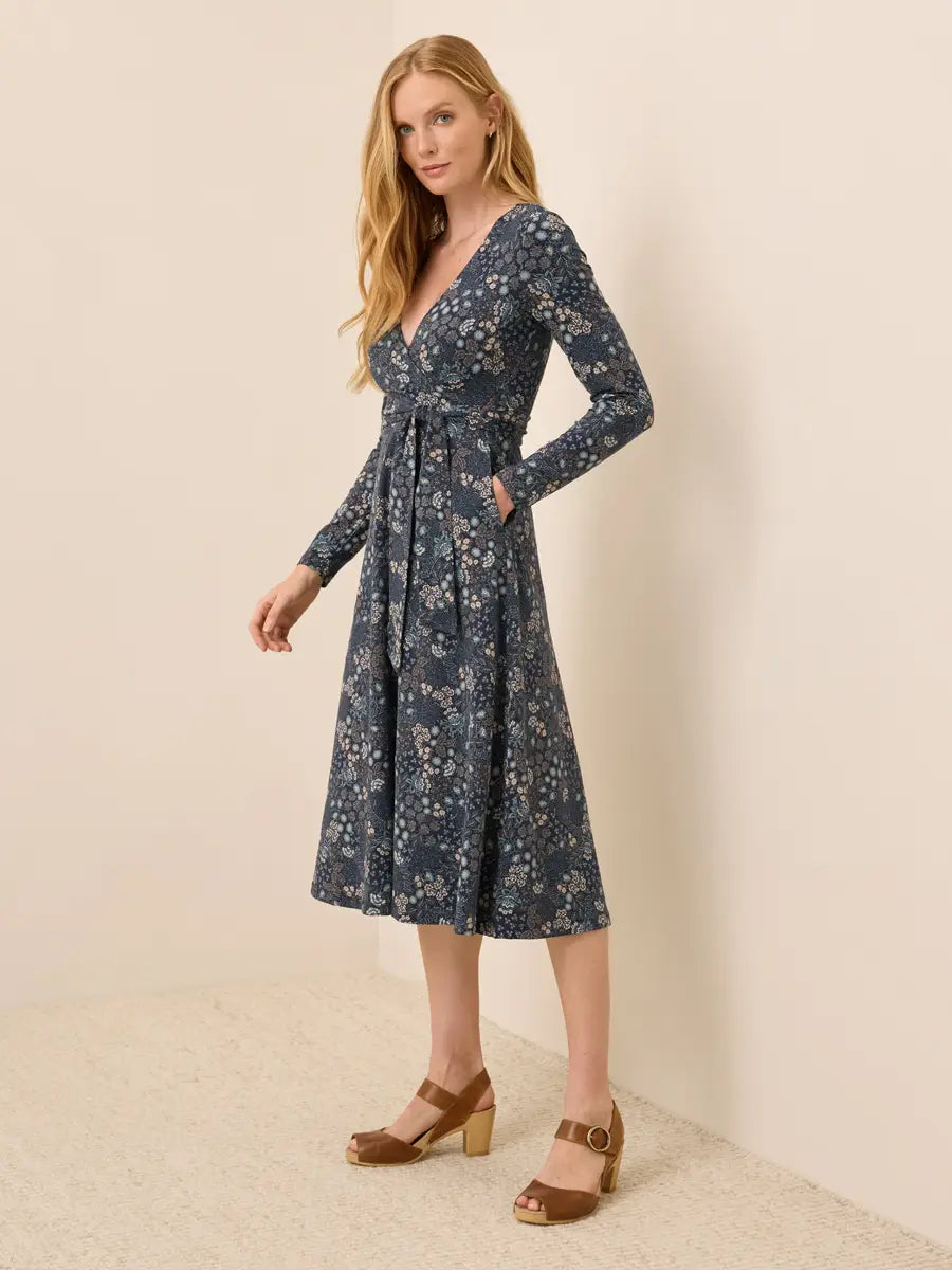 A woman modeling a long-sleeve wrap-style midi dress with a blue floral print, standing against a plain beige wall and textured floor.