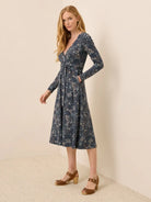 A woman modeling a long-sleeve wrap-style midi dress with a blue floral print, standing against a plain beige wall and textured floor.