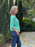A woman with blonde hair stands in profile outdoors on a stone patio, wearing a light mint green long-sleeve top and blue jeans, with tall green ivy-covered hedges behind her.