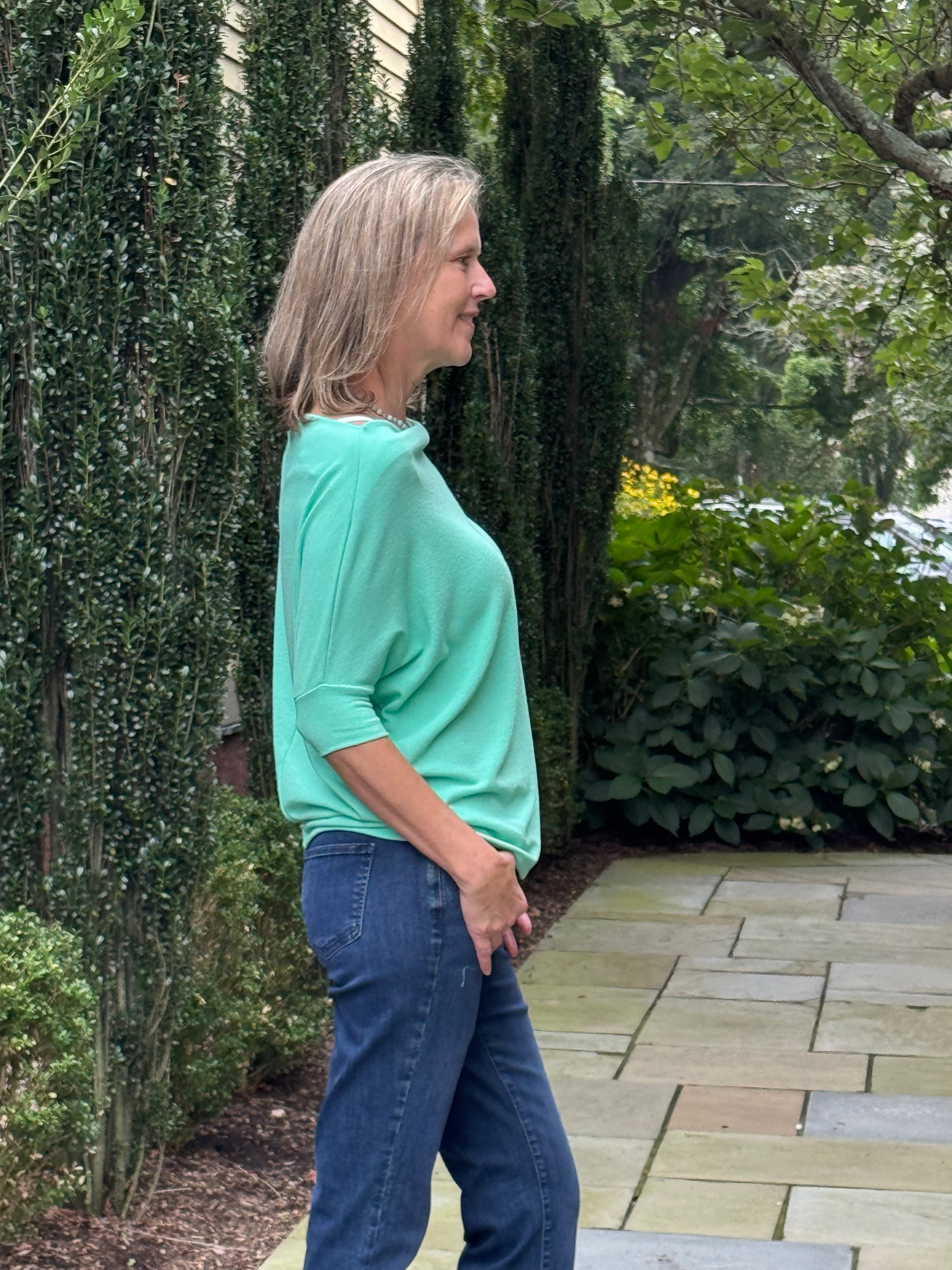A woman with blonde hair stands in profile outdoors on a stone patio, wearing a light mint green long-sleeve top and blue jeans, with tall green ivy-covered hedges behind her.