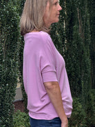 A woman shown in side profile wearing a light pink short-sleeve top and blue jeans, standing outdoors in front of a hedge.