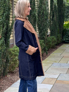 A woman wearing a dark navy blue denim coat with a tan sherpa lining, standing on a stone patio in front of a house with tall green hedges.