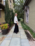 A woman stands on a stone walkway between two houses, wearing a white ruffled blouse and a long black skirt.