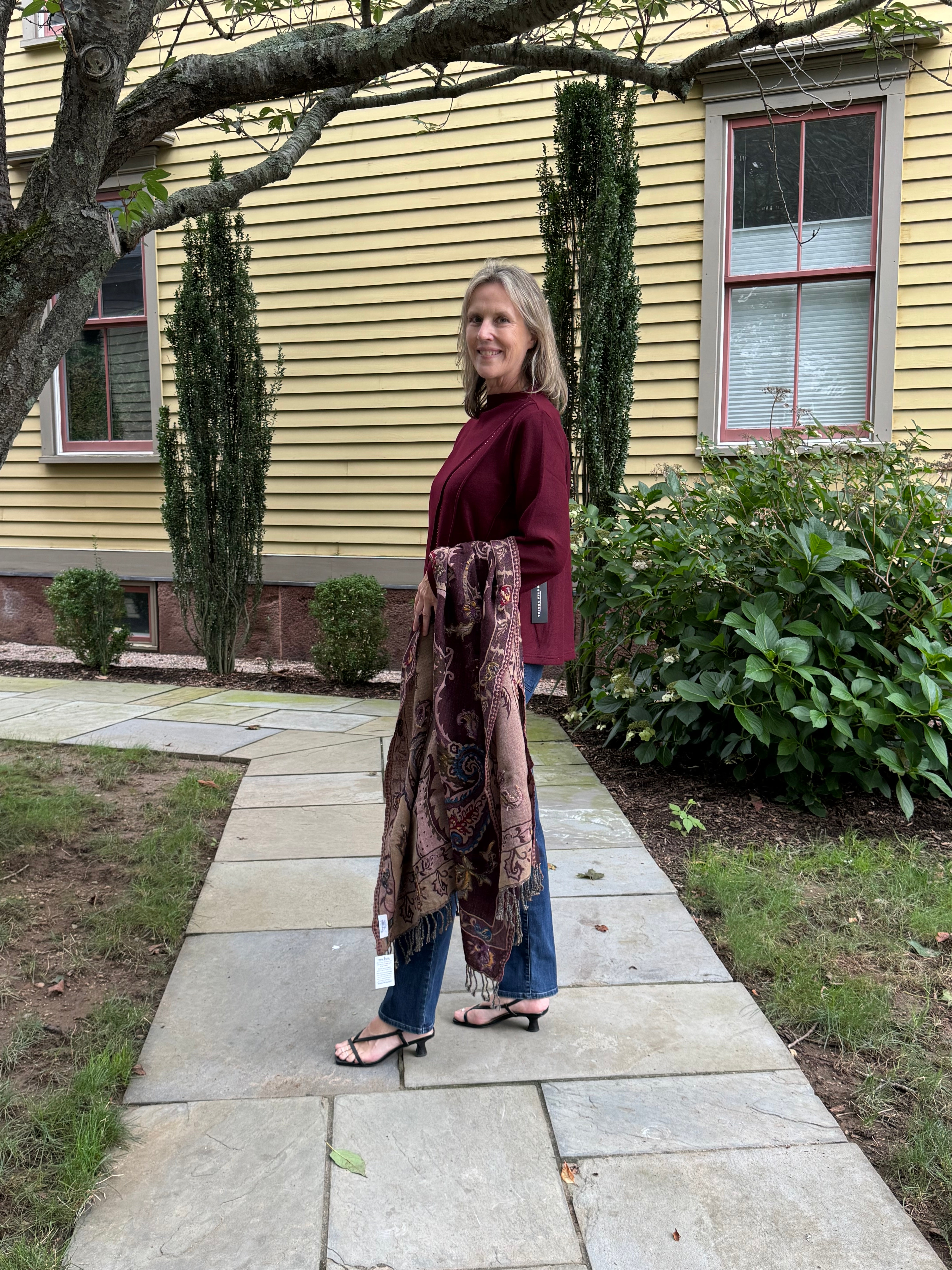 A woman stands on a stone walkway in front of a yellow house, holding up a long patterned scarf or shawl over her arm. She is wearing a burgundy top, blue jeans, and black sandals.