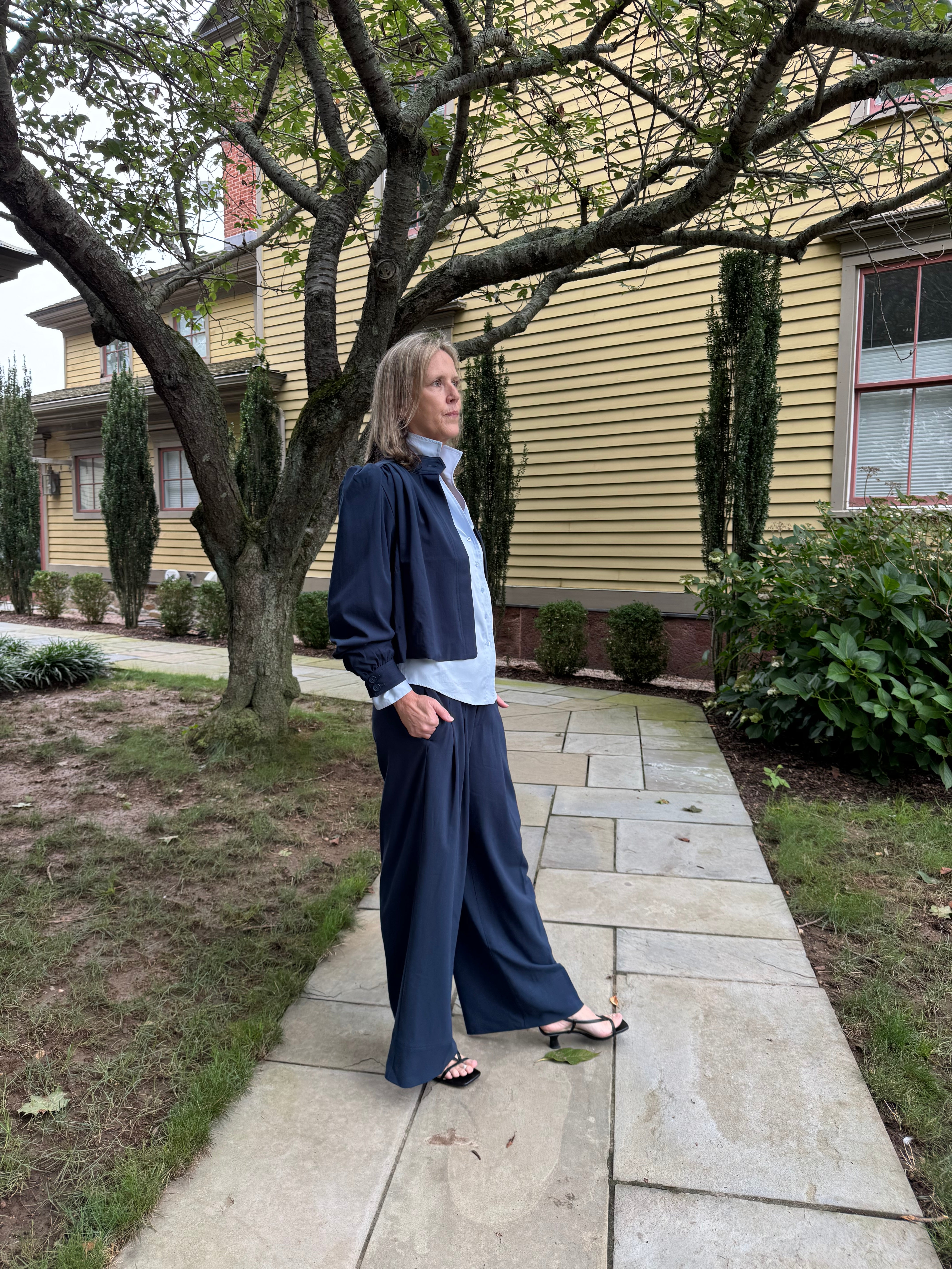 A woman stands on a stone walkway in front of a yellow house, wearing a navy blue wide-leg skirt or pants, a light blue button-up shirt, and black sandals.