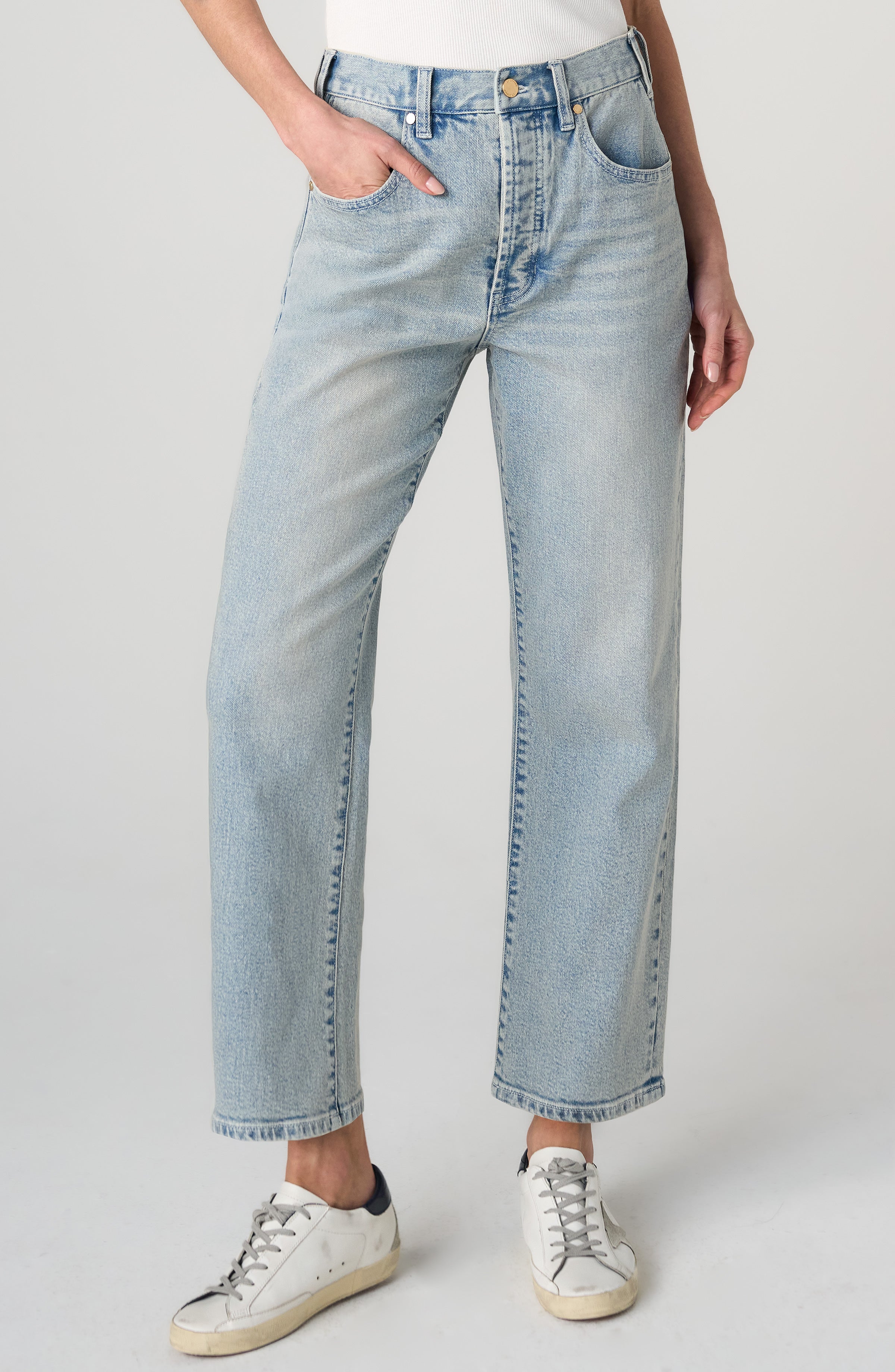 A person wearing light-wash blue straight-leg jeans with a white top and white sneakers, shown from the waist down against a light gray studio background.