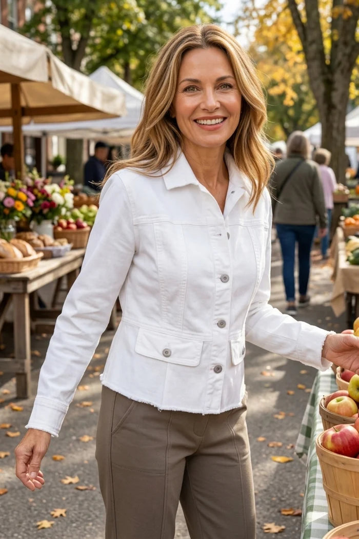 A smiling woman stands at an outdoor farmers market wearing a white cropped button-front jacket with flap pockets and taupe pants. Market stalls with produce and flowers are visible in the background.