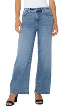 A model wearing a pair of light blue, wide-leg cropped jeans with a faded wash, styled with a white top and navy blue heeled sandals against a plain white background.