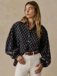Woman wearing a dark patterned blouse and white pants against a beige background