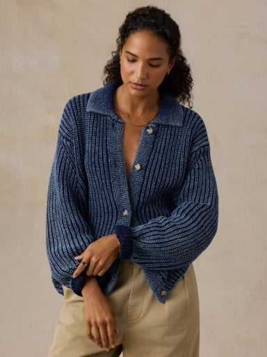 A woman wearing a textured navy blue knit button-up cardigan with voluminous sleeves and beige pants, standing against a neutral textured beige wall.