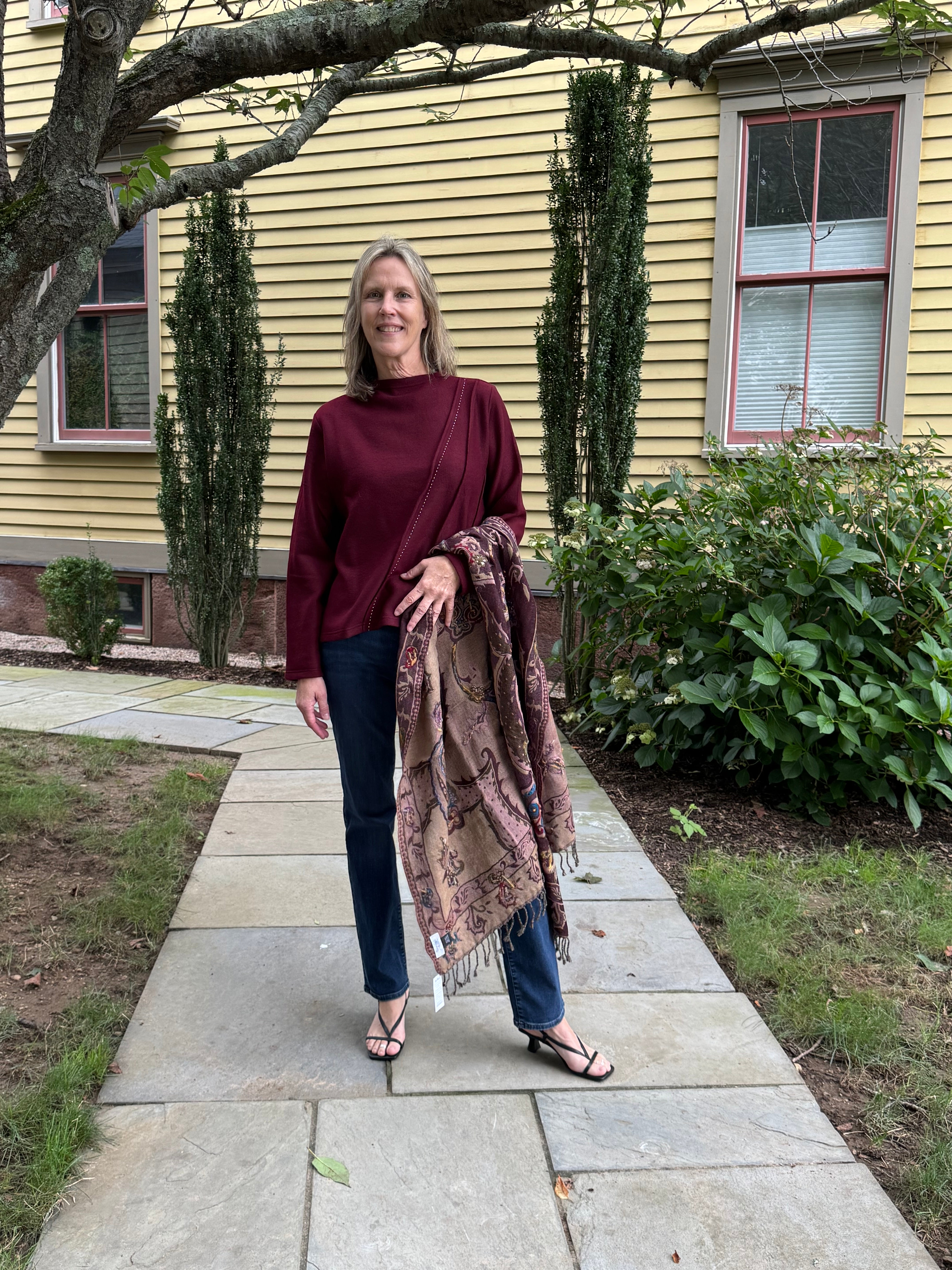 A woman stands on a stone walkway in front of a yellow house, wearing a burgundy mock-neck top, dark blue jeans, and black sandals, while holding a brown and beige patterned shawl.